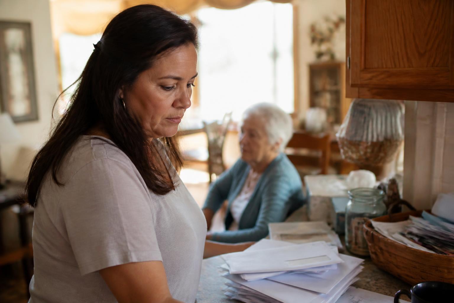 An adult daughter visiting her older mother at home, quietly noticing unopened mail and clutter on the kitchen counter with a concerned expression
